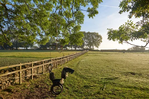 Sunny swing This image, taken in the rural area of the North Yorkshire Moors in the United Kingdom during the autumn of 2018, features a sunny swing as its main subject. Captured in the early morning, the landscape photograph shows a horse-shaped swing made of a tire hanging from the branch of a tree, with sunlight gently illuminating the fields and casting long shadows across the grass. The scene includes a stone wall bordered by a wooden fence stretching out into the distance, with green fields and mature trees under a clear blue sky, encapsulating the peaceful beauty of nature in the region. The image highlights the tranquil and rural character of the North Yorkshire Moors landscape with its open fields and natural charm.